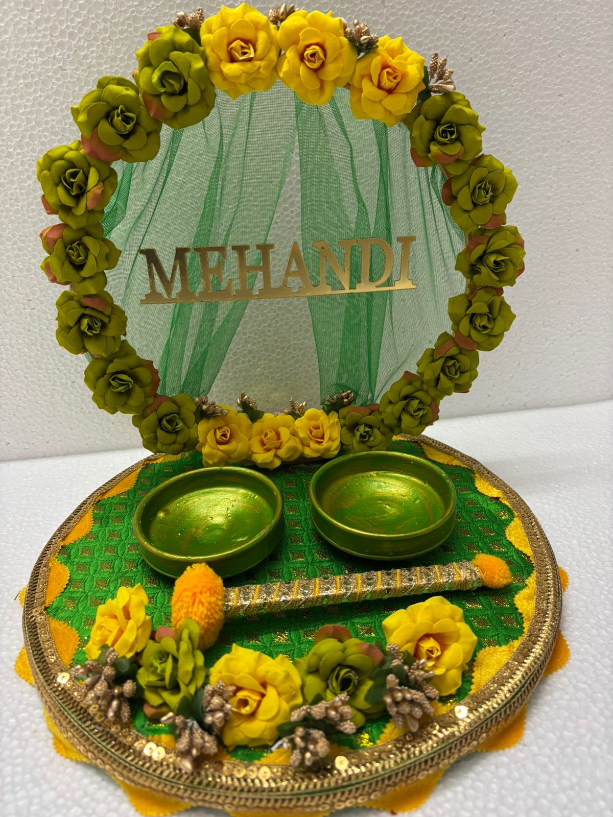 A decorative Mehndi platter featuring a floral design with green and yellow flowers, and a gold-colored structure with the word 'MEHNDI' written in an ornate font. The platter has a green border and is adorned with a golden dish in the center.
