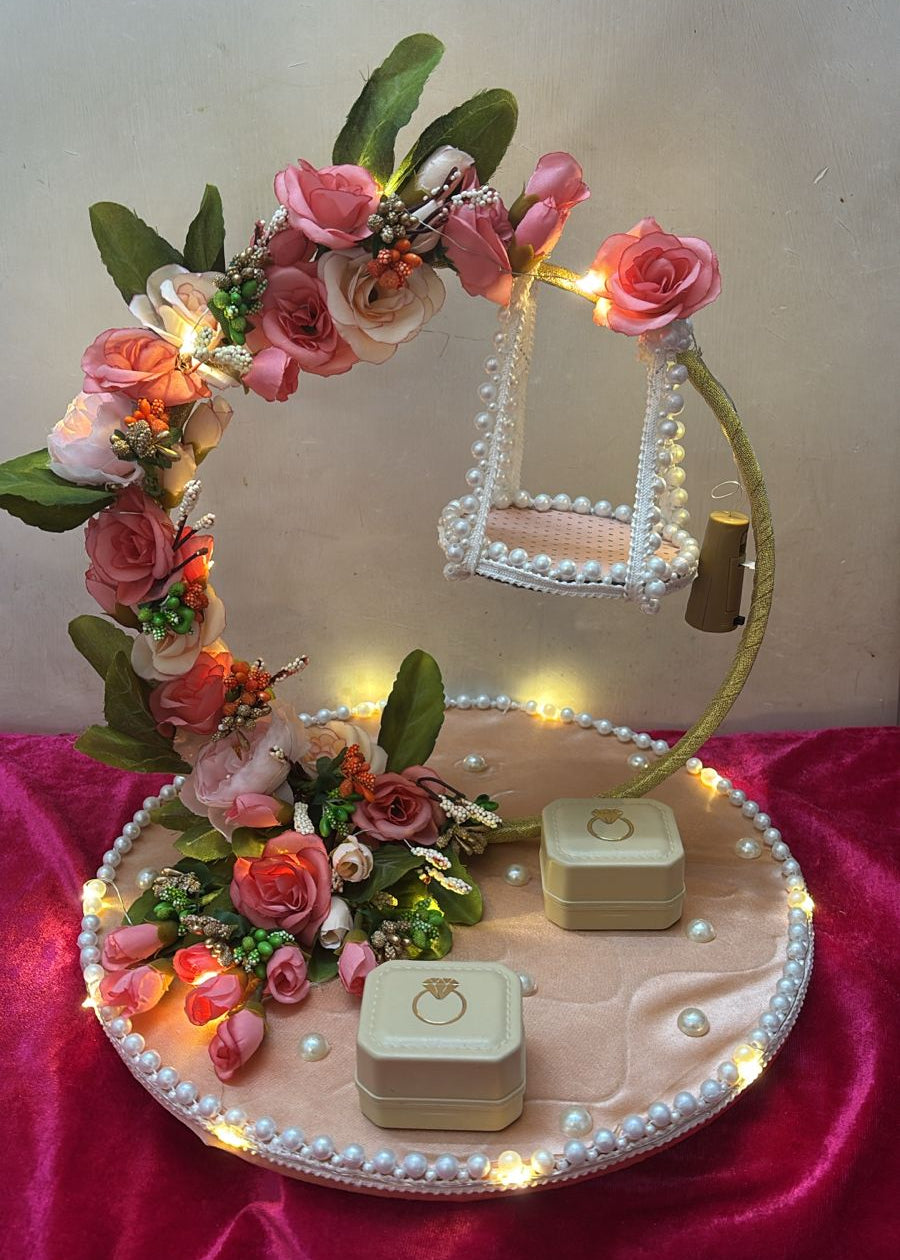 A decorative ring platter with a peach-colored base, adorned with pink and peach flowers, green leaves, and lit by small lights. Two square rings boxes are placed on the platter.
