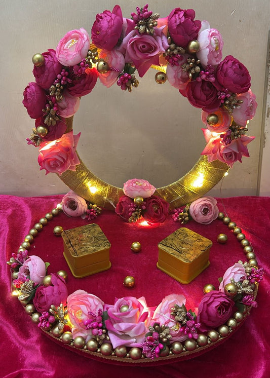 A decorative ring platter with a circular golden tray adorned with pink and purple roses, circular lights, and gold accents, displayed on a vibrant pink fabric.